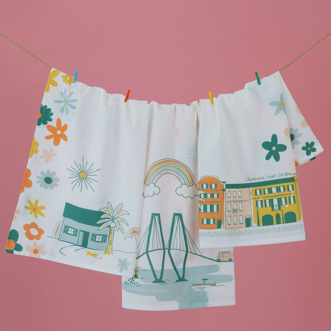 A white fabric curtain panel hanging on a clothesline with colorful clips, featuring a whimsical illustration of a town with a rainbow, buildings, flowers, and a bridge, set against a solid pink background.