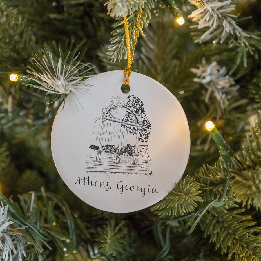 A round white ceramic Christmas ornament with a black line drawing of a birdcage and the text 'Athens, Georgia' hanging from a gold ribbon on a decorated Christmas tree.