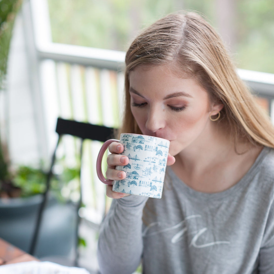 A woman with blonde hair sits at a wooden table, drinking from a white mug with a blue pattern, while reading an open book. She is wearing a gray long-sleeve shirt.