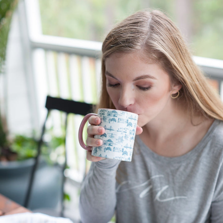 A woman with blonde hair sits at a wooden table, drinking from a white mug with a blue pattern, while reading an open book. She is wearing a gray long-sleeve shirt.